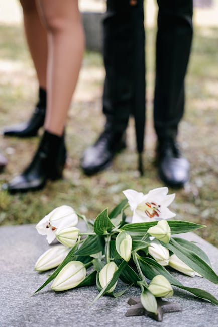 A bouquet of white lilies with large, open blooms and closed buds, featuring long green stems and pointed leaves, carefully arranged and resting on a stone surface. In the background, two people in fo