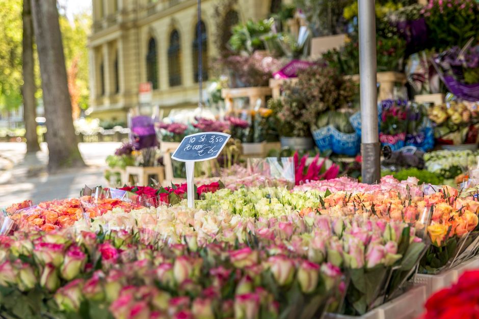 A vibrant outdoor flower market displaying an abundant selection of fresh flowers arranged in bouquets and loose stems. Prominently featured are roses in shades of pink, peach, and cream, with some pi