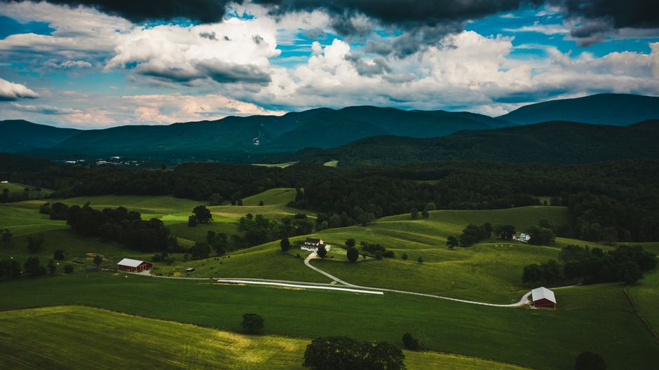 The image depicts a scenic rural landscape with rolling green fields dotted with a few small white and red farm buildings. A winding dirt road leads through the fields, set against a backdrop of dense